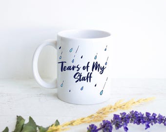 Op de afbeelding: Een witte keramische mok met de donkerblauwe tekst "Tears of My Staff" op de voorzijde gedrukt. Het ontwerp bevat verspreide lichtblauwe en donkerblauwe regendruppelillustraties rondom de tekst, met een gebogen handvat aan de linkerkant.