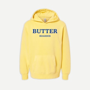 Puede incluir: Una sudadera con capucha de color amarillo pálido que presenta un bolsillo tipo canguro frontal y el texto BUTTER HOARDER impreso en letras mayúsculas de color azul intenso en el pecho. Tiene mangas largas, puños acanalados y capucha.
