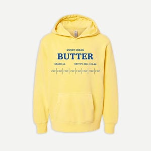 May include: A bright yellow hooded sweatshirt featuring blue text that reads SWEET CREAM BUTTER, GRADE AA, NET WT. 4OZ. (113.4g), and a measurement scale from 1 TBSP to 8 TBSP. The garment includes a front kangaroo pocket and long sleeves.