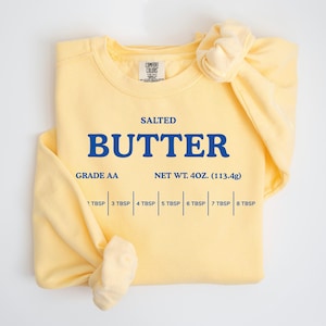 May include: A pale yellow crewneck sweatshirt featuring blue screen-printed text that reads SALTED BUTTER, GRADE AA, NET WT. 4OZ. (113.4g), and a measurement scale from 2 TBSP to 8 TBSP. The garment has a Comfort Colors brand neck label.