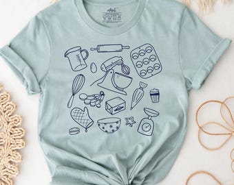 May include: A light sage green t-shirt featuring a dark blue line art graphic of various baking tools including a measuring cup, rolling pin, muffin tin, whisk, stand mixer, piping bag, oven mitt, mixing bowl, and a kitchen scale.