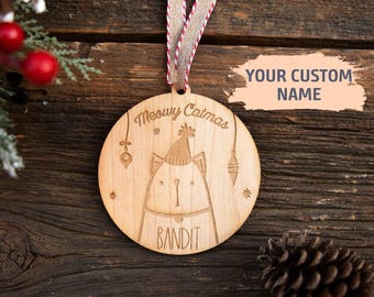 May include: A round wooden ornament featuring an engraved cat wearing a party hat, the text Meowy Catmas, and the name Bandit. The ornament hangs from a red and white striped ribbon. A graphic overlay displays the text YOUR CUSTOM NAME.