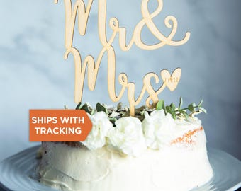 May include: A light-colored wooden cake topper featuring the cursive text Mr & Mrs with a small heart shape containing the date 7.19.22. The topper is positioned on top of a round white frosted cake decorated with white flowers and green leaves.