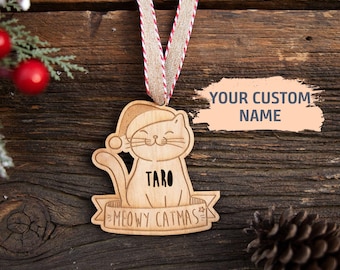May include: A light-colored wooden ornament shaped like a cat wearing a Santa hat. The cat has the name Taro engraved on its chest and sits above a banner with the text MEOWY CATMAS. It hangs from a beige ribbon with red and white edges.