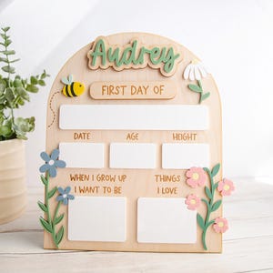 Peut inclure: Un panneau en bois arqué avec le nom Audrey en vert, portant le texte FIRST DAY OF, DATE, AGE, HEIGHT, WHEN I GROW UP I WANT TO BE et THINGS I LOVE. Il comprend des espaces rectangulaires blancs pour écrire, une abeille jaune et des fleurs.