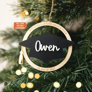 Peut inclure: Ornement de graduation en bois avec un mortier noir et le nom "Owen" en écriture blanche. L'ornement circulaire porte les inscriptions "Westfield Junior High School" et "Class of 2023". Une gland doré est suspendu.