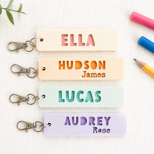 May include: Four rectangular keychains in pastel colours, each featuring a silver metal lobster clasp. The keychains display the names ELLA, HUDSON James, LUCAS, and AUDREY Rose, with the primary names cut out to reveal the colour underneath.