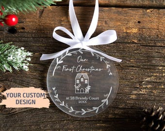 Pode incluir: Um enfeite de Natal redondo em acrílico transparente com um laço de fita branca no topo. O enfeite é gravado com um design de guirlanda, uma ilustração de casa e o texto Our First Christmas at 28 Brandy Court 20XX em letras brancas.