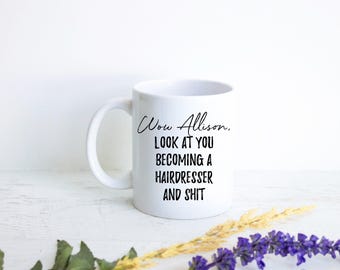 Peut inclure: Un mug en céramique blanche avec une anse sur le côté gauche. L'avant du mug présente un texte imprimé en noir qui dit : Wow Allison, LOOK AT YOU BECOMING A HAIRDRESSER AND SHIT. Le mug a une finition lisse et brillante et une forme cylindrique.