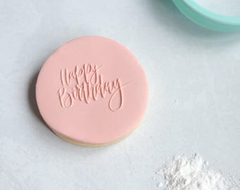 May include: A round cookie covered in smooth light pink fondant with the words Happy Birthday embossed in a cursive script font. A small pile of white powdered sugar sits nearby, and a light blue circular object is visible in the corner.