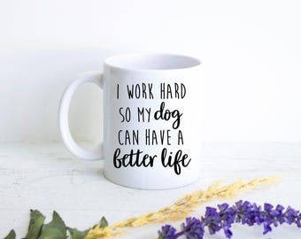 May include: A white ceramic mug with a handle on the left side. The mug features black printed text that reads: I WORK HARD SO MY dog CAN HAVE A better life. The font style alternates between a clean sans-serif and a cursive script typeface.