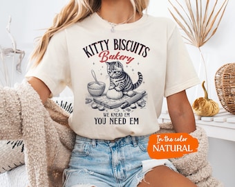 Può includere: Una t-shirt color naturale con la grafica di un gattino tigrato che impasta la pasta insieme a una ciotola, un mattarello e farina. Il testo recita: KITTY BISCUITS Bakery WE KNEAD EM YOU NEED EM. Abbinata a pantaloncini di jeans azzurri.