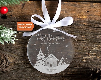 Pode incluir: Um ornamento circular de acrílico transparente com um laço de fita branca. O design apresenta uma cena de inverno gravada em branco com uma casa, árvores e flocos de neve. O texto diz: First Christmas at 124 Privet Drive, The Millers, 2020.