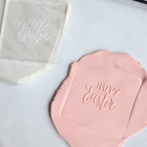May include: A square clear acrylic stamp featuring the embossed text Happy Easter in a cursive script font, positioned next to a piece of light pink modeling clay that has been imprinted with the same Happy Easter design in a square shape.