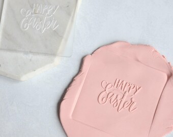 May include: A square clear acrylic stamp featuring the embossed text Happy Easter in a cursive script font, positioned next to a piece of light pink modeling clay that has been imprinted with the same Happy Easter design in a square shape.