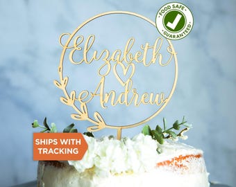Puede incluir: Un adorno circular de madera para pastel con los nombres Elizabeth y Andrew en letra cursiva y un pequeño corazón en el centro. El diseño está enmarcado por un fino anillo de madera con detalles de hojas a la izquierda y un soporte.