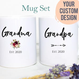 May include: Two white ceramic mugs featuring black text and graphics. The left mug displays the word Grandma, a floral bouquet, and EST. 2020. The right mug displays the word Grandpa, an arrow graphic, and EST. 2020. Text above reads Mug Set and Your Custom Design.