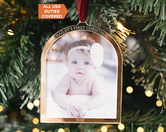 May include: A gold-toned arched metal Christmas ornament featuring a photograph of a baby wearing a white headband. The ornament is engraved with the text HAILEY'S FIRST CHRISTMAS at the top and 2025 at the bottom, suspended by a dark red ribbon.