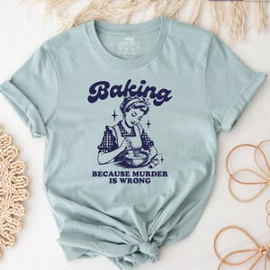 Könnte beinhalten: Ein hellblaues kurzärmeliges T-Shirt mit einer dunkelblauen Grafik einer Person in einer Schürze, die in einer Schüssel rührt. Der Text darüber lautet Baking und der Text darunter lautet BECAUSE MURDER IS WRONG. Rundhalsausschnitt und umgeschlagene Ärmel.