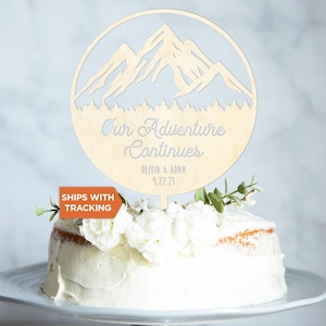 May include: A circular light-colored wooden cake topper featuring a laser-cut mountain range and forest silhouette. The text reads Our Adventure Continues, Olivia & Adam, and 9.22.21. The topper is inserted into a white frosted cake with flowers.
