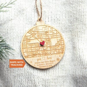 May include: A round wooden ornament featuring an engraved street map with a small red heart placed at a specific location. The text reads "jen & ben", "Where Our Story Began", "36.25413 N, 115.2424 W", and "November 3, 2021" with star accents.