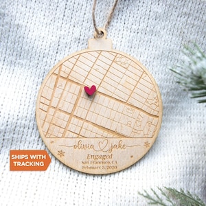 May include: Wooden Christmas ornament with a map design, a red heart, and the names "Olivia & Jake" engraved. The ornament also includes the text "Engaged San Francisco, CA February 3, 2020". It is hanging by a twine loop.