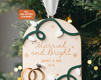 May include: A light-coloured wooden arch-shaped ornament featuring the text Married and Bright, the names GRANT & AVA, and the date 5.3.22. It is decorated with green wire, white light bulb shapes, gold snowflake icons, and two gold rings.