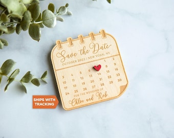 Puede incluir: Una placa de madera clara con forma de calendario que muestra el texto Save the Date, October 2022, New York, NY, y For the wedding of Chloe and Rob. Un pequeño corazón de madera roja está colocado sobre el número 8 del calendario.