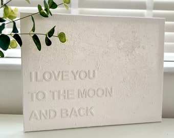 May include: White textured canvas sign with the words "I love you to the moon and back" embossed in white.
