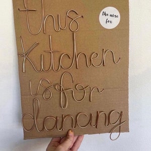 Puede incluir: Un letrero de alambre de cobre sobre cartón. El letrero dice "this kitchen is for dancing" en escritura cursiva. Una pequeña pegatina blanca con el texto "the wire fox" está en la esquina superior derecha.