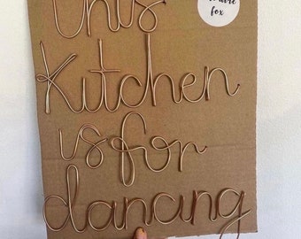 May include: A copper wire sign on a cardboard backing. The sign reads "this kitchen is for dancing" in cursive script. A small white sticker with the text "the wire fox" is in the upper right corner.