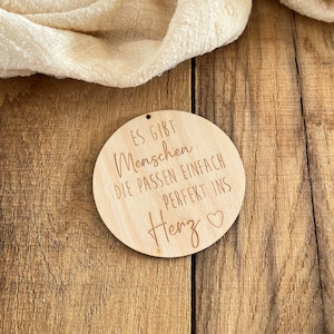 Könnte beinhalten: Ein rundes Holzschild mit dem Text "Es gibt Menschen die passen einfach perfekt ins Herz" mit einem kleinen Herzsymbol.