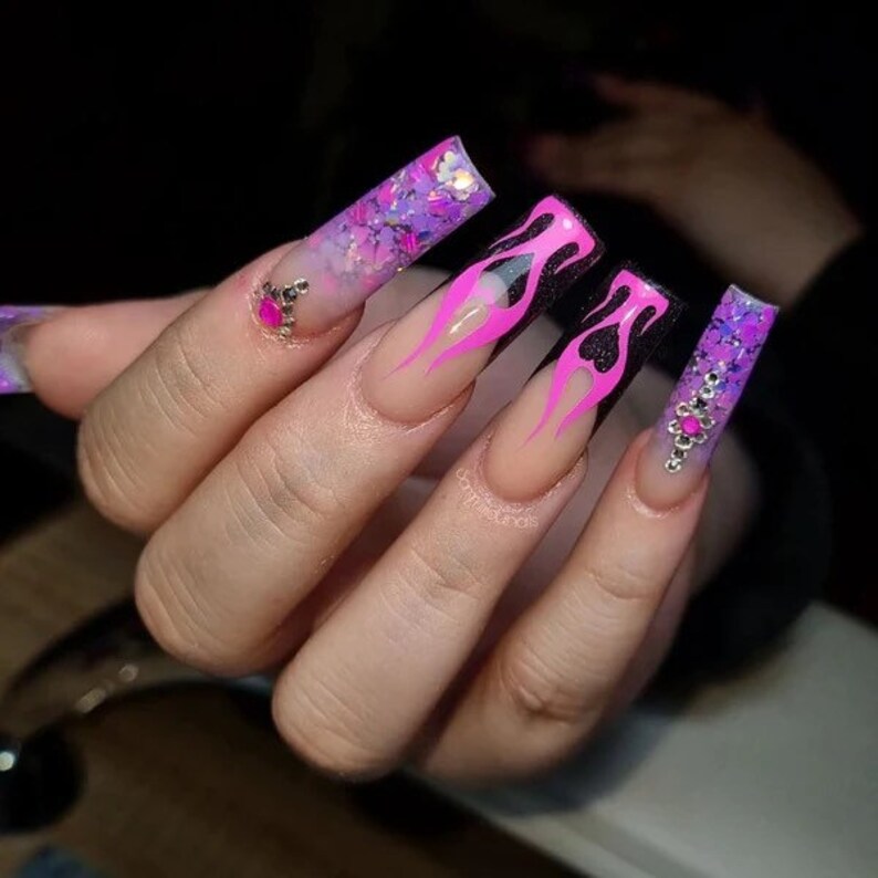 Puede incluir: U&ntilde;as acr&iacute;licas largas de color nude con un dise&ntilde;o de llamas negras y rosas. Algunas u&ntilde;as tienen un acabado brillante y pedrer&iacute;a.