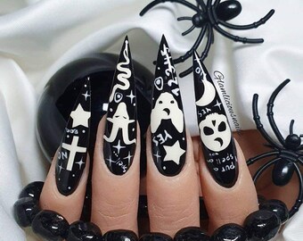 Puede incluir: Uñas de Halloween en blanco y negro con diseños espeluznantes. Cada uña presenta un diseño diferente, incluyendo una cruz, un fantasma, una serpiente y una calavera. Las uñas son largas y puntiagudas, con un acabado brillante. Hay arañas negras decorativas y una esfera negra en el fondo.