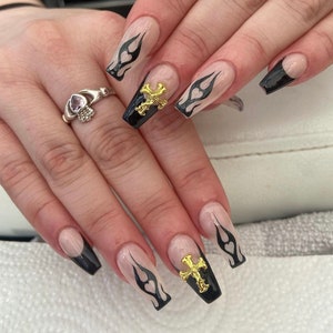 Puede incluir: Primer plano de una mano con u&ntilde;as largas de color nude con puntas negras y dise&ntilde;os de llamas. Algunas u&ntilde;as tienen detalles de cruces dorados.