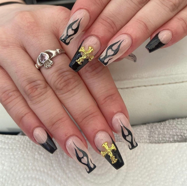 Puede incluir: Primer plano de una mano con u&ntilde;as largas de color nude con puntas negras y dise&ntilde;os de llamas. Algunas u&ntilde;as tienen detalles de cruces dorados.