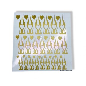 Puede incluir: Una hoja de pegatinas doradas para u&ntilde;as con un dise&ntilde;o de llama y un recorte en forma de coraz&oacute;n. Hay 24 pegatinas en la hoja.