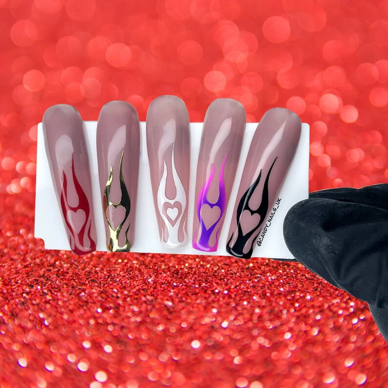 Puede incluir: Cinco u&ntilde;as postizas largas en forma de almendra con dise&ntilde;os de llamas de diferentes colores y un coraz&oacute;n en la punta. Los colores son rojo, dorado, blanco, morado y negro.
