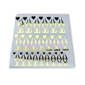 Puede incluir: Una hoja de pegatinas para u&ntilde;as doradas y negras con un dise&ntilde;o de llama y un coraz&oacute;n recortado. Las pegatinas est&aacute;n en una hoja de respaldo blanca.
