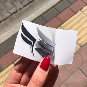 Może przedstawiać: Zestaw naklejek w kształcie skrzydeł w kolorze czarnym i srebrnym, częściowo odklejonych od białego podłoża. Naklejki mają warstwowy wz&oacute;r, z czarnymi skrzydłami po lewej i srebrnymi po prawej stronie. Tło to mieszanka chodnika i światła słonecznego.