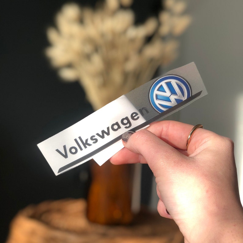 Może przedstawiać: Prostokątna naklejka z logo Volkswagena i nazwą marki. Logo to niebiesko-biały emblemat, a słowo "Volkswagen" jest wydrukowane na czarno. Naklejka jest trzymana w dłoni.