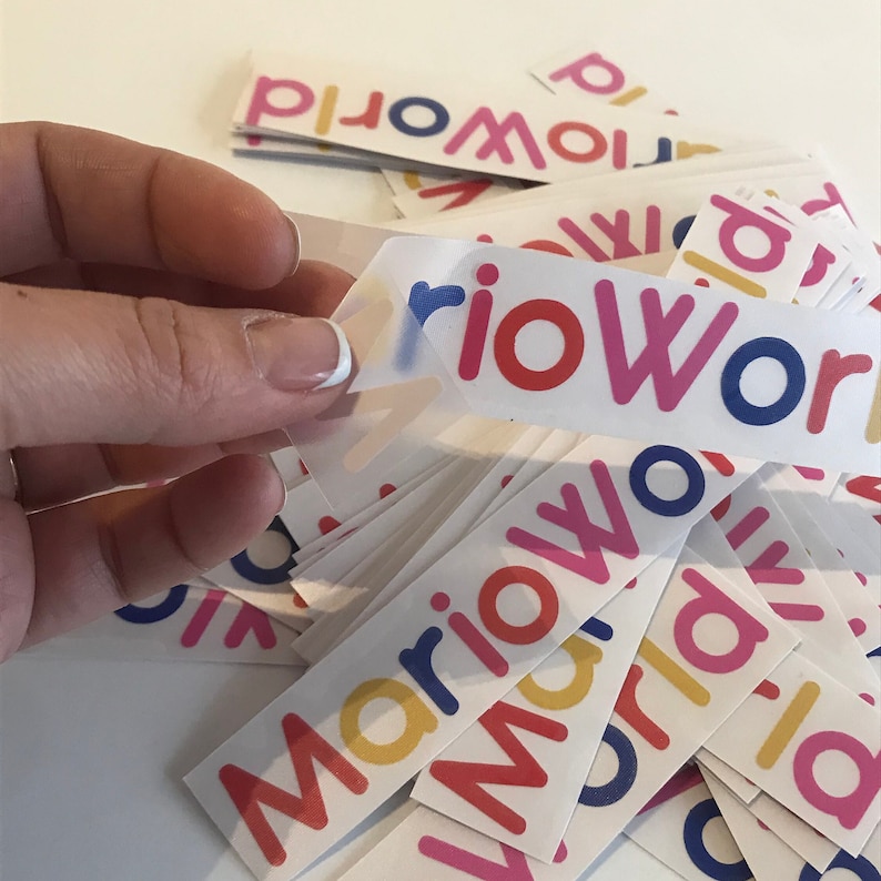 Może przedstawiać: Ręka trzyma naklejkę z napisem "Mario World" w kolorowych literach. Naklejki są białe z tekstem w kolorze czerwonym, r&oacute;żowym, ż&oacute;łtym, niebieskim i pomarańczowym. W tle znajduje się stos podobnych naklejek.