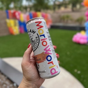 Może przedstawiać: Biała puszka hard seltzer z napisem "MARIO WORLD" w kolorowych literach. Na puszce widnieje r&oacute;wnież napis "White Claw Hard Seltzer Ruby Grapefruit" i ma krople wody na powierzchni. Tło to zielony trawnik.