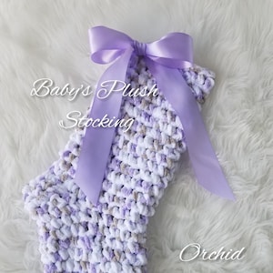 May include: A purple and white plush stocking with a lavender bow. The stocking has the text "Baby's Plush Stocking" and "Orchid and Bee's" on it.