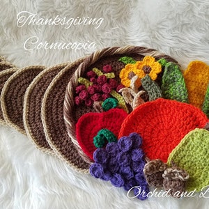 May include: A crocheted cornucopia with a brown and beige base, filled with crocheted fruits, vegetables, and flowers in shades of red, orange, yellow, green, and purple. The cornucopia is decorated with the text "Thanksgiving Cornucopia" and the signature "Orchid and Bee's".