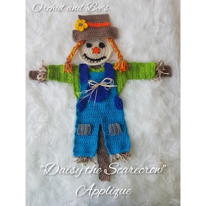 May include: A crocheted scarecrow applique with a brown hat, orange hair, green shirt, blue overalls, and a brown stick. The applique is labeled "Daisy the Scarecrow" and "Applique".