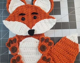 Puede incluir: Un juguete de zorro de crochet con piel naranja y blanca, ojos marrones y nariz negra. El zorro tiene cuatro huellas de patas marrones y una cola larga naranja.