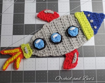 May include: A crocheted rocket toy with a grey body, red and yellow flames, and blue and yellow details. The rocket has the text "Rocketeering Rocket" and "Orchid and Bee's" on it.
