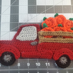 May include: A crocheted red pickup truck carrying a load of orange pumpkins with green stems. The truck has gray windows and wheels, and a brown bed. A fall-themed decorative item.