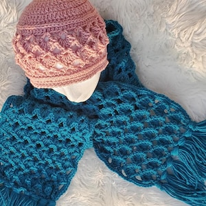 May include: A pink crocheted beanie with a blue crocheted scarf with tassels. The scarf and beanie are made with a lacy stitch.